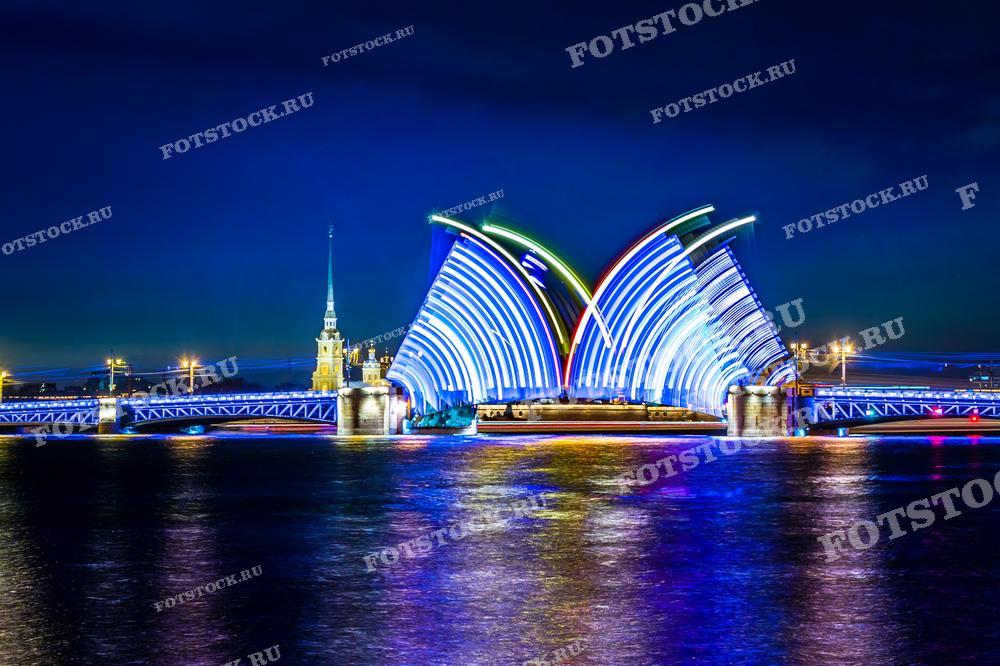 На фотографии изображен ночной вид Дворцового моста в Санкт-Петербурге во время его разводки. Яркая подсветка моста сияет множеством оттенков синего цвета, создавая живописный силуэт против тёмного ночного неба. Петропавловская крепость на заднем плане добавляет узнаваемости изображению. Фотография подойдёт для использования в материалах о достопримечательностях Санкт-Петербурга, туристических буклетах, рекламе ночной жизни города и публикациях об искусстве уличной фотографии.